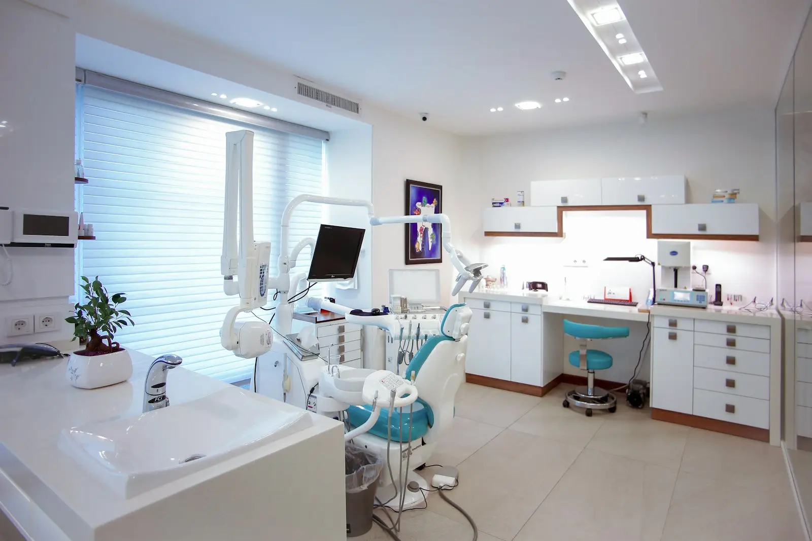 A bright dental surgery with a teal dental chair, a sink counter and white cabinets